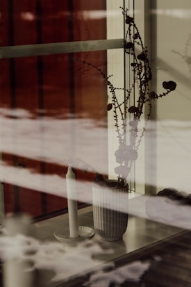 A still life scene featuring a tall, fluted ceramic vase with dried branches and flowers on a windowsill. Next to the vase is a simple white candle on a small plate, all behind a glass window with the outside view slightly reflected. The reflection and muted tones create a serene and contemplative atmosphere.