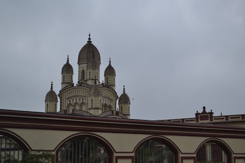 A grand temple structure featuring multiple intricately designed domes, bordered by a decorative wall with arch designs. The architecture includes ornate details and a blend of traditional and classical styles under a cloudy sky.