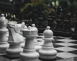 A giant outdoor chess set with large white and black chess pieces arranged on a black and white checkered board. The pieces include a knight, rook, bishop, and pawns. The background features lush green foliage and some flowers, adding to the outdoor garden setting.