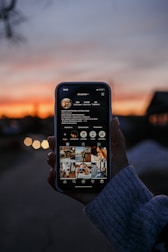 A hand is holding a smartphone displaying a social media profile with multiple photos on the screen. The background features a sunset with a gradient sky transitioning from orange to blue, creating a serene atmosphere.