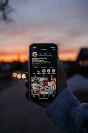 A hand is holding a smartphone displaying a social media profile with multiple photos on the screen. The background features a sunset with a gradient sky transitioning from orange to blue, creating a serene atmosphere.
