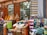 A bustling market scene with vibrant textiles and mounds of colorful spices displayed in large cones outside a shop with wooden doors. A person is visible, possibly a vendor, amidst the display of goods, which includes neatly folded rugs in various patterns and colors.