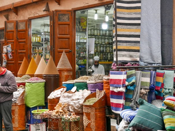 A bustling market scene with vibrant textiles and mounds of colorful spices displayed in large cones outside a shop with wooden doors. A person is visible, possibly a vendor, amidst the display of goods, which includes neatly folded rugs in various patterns and colors.