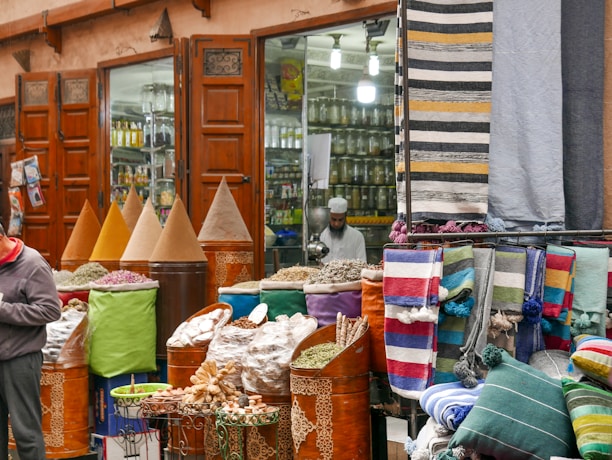 A bustling market scene with vibrant textiles and mounds of colorful spices displayed in large cones outside a shop with wooden doors. A person is visible, possibly a vendor, amidst the display of goods, which includes neatly folded rugs in various patterns and colors.