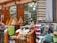 A bustling market scene with vibrant textiles and mounds of colorful spices displayed in large cones outside a shop with wooden doors. A person is visible, possibly a vendor, amidst the display of goods, which includes neatly folded rugs in various patterns and colors.