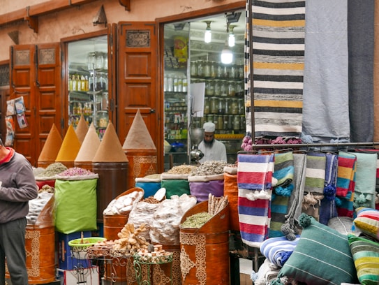 A bustling market scene with vibrant textiles and mounds of colorful spices displayed in large cones outside a shop with wooden doors. A person is visible, possibly a vendor, amidst the display of goods, which includes neatly folded rugs in various patterns and colors.