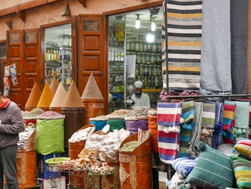 A bustling market scene with vibrant textiles and mounds of colorful spices displayed in large cones outside a shop with wooden doors. A person is visible, possibly a vendor, amidst the display of goods, which includes neatly folded rugs in various patterns and colors.