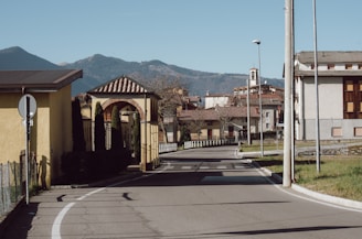 A quiet suburban street curves gently to the left, with houses featuring sloped roofs and white and yellow walls lining the road. A small bell tower is visible in the distance against a backdrop of hills under a clear blue sky. Well-maintained shrubbery and trees provide some greenery along the sidewalk.