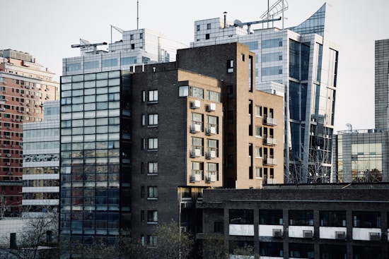 A cluster of modern office buildings with reflective glass facades and unique architectural designs, featuring a prominent brown brick structure in the foreground. The scene includes a mix of straight lines and angular shapes, creating a sense of urban complexity.