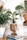 A cozy room corner features several potted plants, including a large woven planter with a lush green plant. A small statue of a meditative figure sits atop a stack of books on the floor. The light-colored wooden floor and a nearby table lamp add to the serene atmosphere.