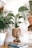 A cozy room corner features several potted plants, including a large woven planter with a lush green plant. A small statue of a meditative figure sits atop a stack of books on the floor. The light-colored wooden floor and a nearby table lamp add to the serene atmosphere.