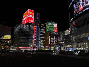 A bustling cityscape at night featuring numerous brightly lit buildings adorned with colorful signage. High-rise structures display prominent advertisements in Japanese, with a mix of digital screens and neon lights. The street is lively with cars and pedestrians, contributing to an urban atmosphere.