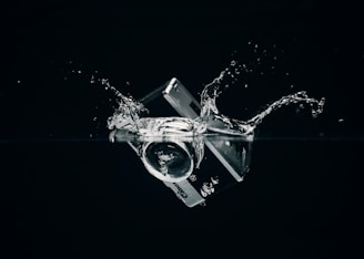 A credit card and camera lens are submerged in water, creating splashes against a dark background.