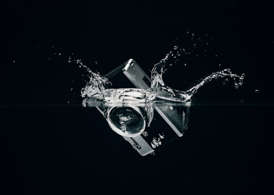 A credit card and camera lens are submerged in water, creating splashes against a dark background.