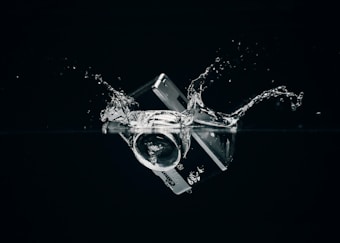 A credit card and camera lens are submerged in water, creating splashes against a dark background.