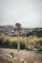 A wooden signpost stands amidst a natural landscape featuring grassy terrain and rocky mountains in the background. The sky is overcast with thick cloud cover, suggesting a cool and misty atmosphere. The signpost has red markings and directs towards different hiking paths, creating a sense of adventure and exploration.