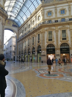 An ornate indoor shopping gallery with a high arched glass ceiling and detailed classical architecture. Marble floors feature intricate patterns, while luxury storefronts like Prada are visible. Several people walk, enjoying the brightly lit, elegant environment.