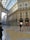 An ornate indoor shopping gallery with a high arched glass ceiling and detailed classical architecture. Marble floors feature intricate patterns, while luxury storefronts like Prada are visible. Several people walk, enjoying the brightly lit, elegant environment.