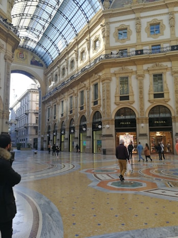 An ornate indoor shopping gallery with a high arched glass ceiling and detailed classical architecture. Marble floors feature intricate patterns, while luxury storefronts like Prada are visible. Several people walk, enjoying the brightly lit, elegant environment.