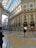 An ornate indoor shopping gallery with a high arched glass ceiling and detailed classical architecture. Marble floors feature intricate patterns, while luxury storefronts like Prada are visible. Several people walk, enjoying the brightly lit, elegant environment.
