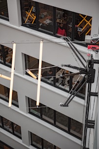 Several floors of an office building are visible, with large windows allowing a view into multiple offices. Inside the offices, people are seen working at desks with computers, and various office items are scattered around. In front of the building's facade, there is a large, metallic structure with illuminated, elongated tubes hanging from it.