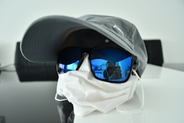 A gray baseball cap and blue-tinted sunglasses are placed on top of a white face mask, which is situated on a reflective surface. The sunglasses reflect an image of a person taking a photo, and the background is minimalistic with a dark object in the distance.