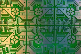 Complex and detailed circuit board featuring symmetrical patterns and intricate pathways. The design includes a repeating geometrical motif with rounded and linear elements, embedded with multiple holes for components.