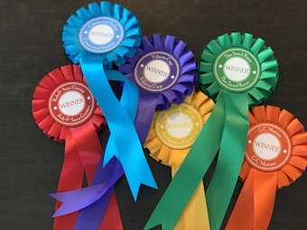 A collection of colorful award ribbons, each labeled as a winner. The ribbons are of various colors including red, blue, purple, green, yellow, and orange, arranged on a dark surface. Each ribbon is marked with different event names such as 'Conaccord Stakes' and 'The Sand Race'.