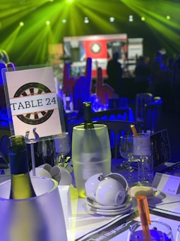 A table set for an event, featuring a sign labeled 'Table 24' with the backdrop of a dartboard design. The table includes a wine bottle in a cooler, stacked white teacups, wine glasses, and a clipping with the text 'White Collar Darts 2020.' The background is dimly lit with colorful lighting, creating a vibrant atmosphere.