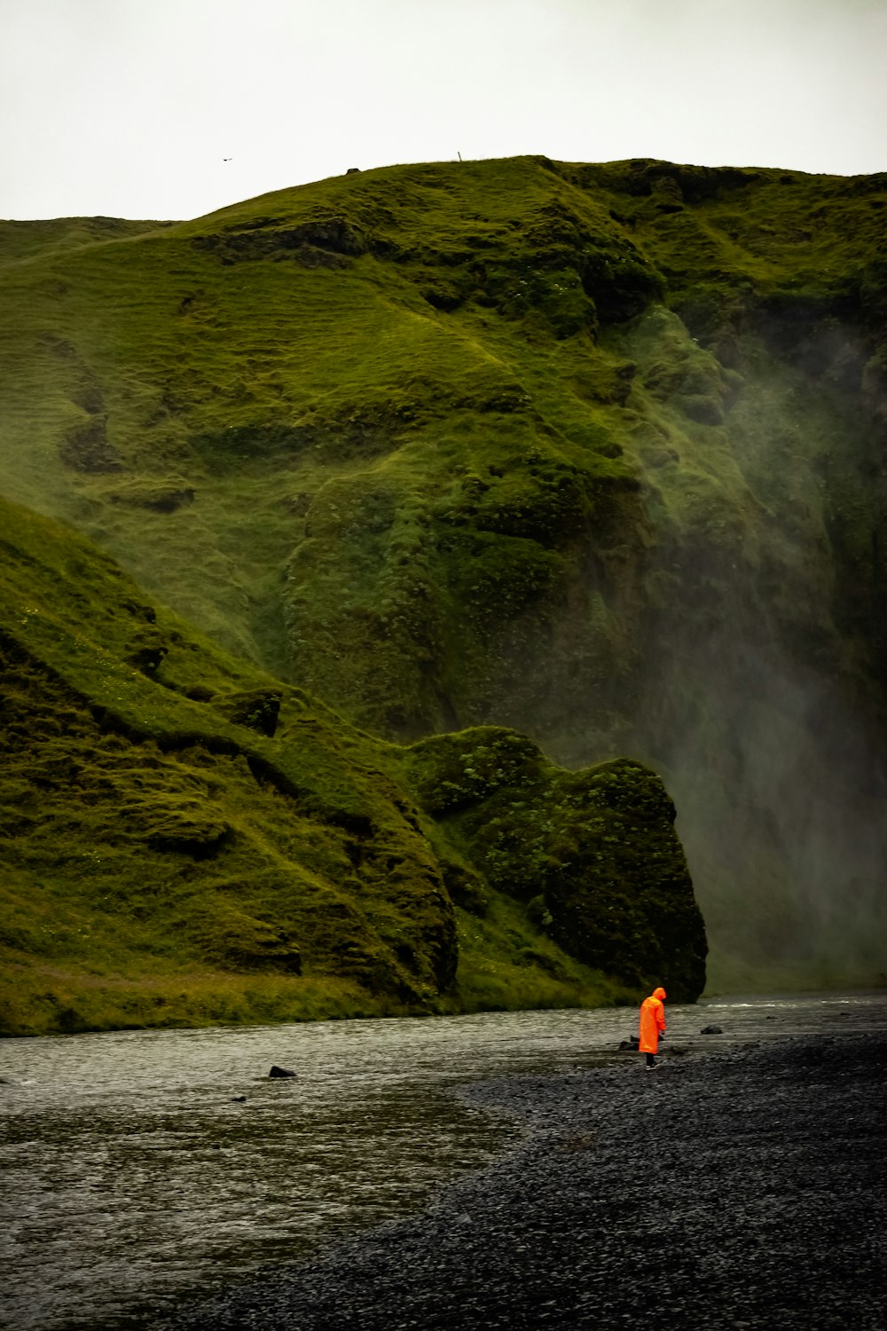 person in orange jacket standing on water near green mountain during daytime