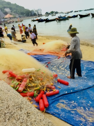A group of people gather on a sandy beach engaged in fishing-related activities. Fishing nets, strewn on the blue tarp, are prominent, with several red floats attached. Small fish scatter across the surface around the nets. In the background, traditional boats float on the calm water, and a green, hilly landscape rises with several buildings overlooking the shore.