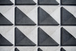 A grid of three-dimensional square tiles with triangular patterns, each tile casting shadows that create a sense of depth. The tiles have a geometric design and are arranged in a repeating pattern.