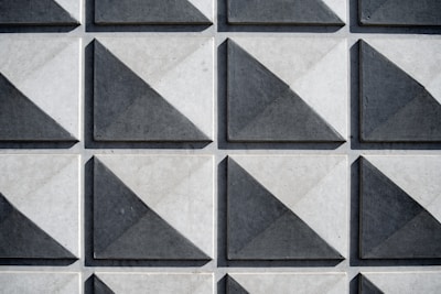 A grid of three-dimensional square tiles with triangular patterns, each tile casting shadows that create a sense of depth. The tiles have a geometric design and are arranged in a repeating pattern.