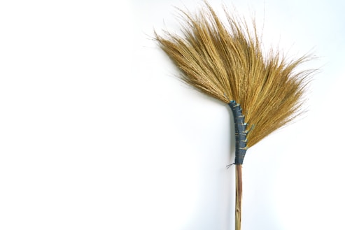 A hand broom made from natural materials, with a fan shape consisting of straw-like bristles. The bristles are tightly bound by a blue fabric wrap near the handle, which is made of wood. The background is a plain white surface.