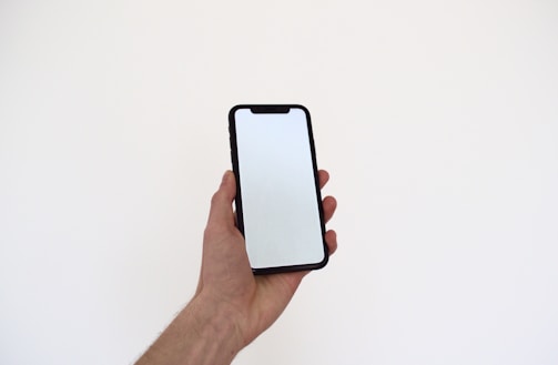 A hand holding a smartphone with a black casing. The screen is blank and reflects light, indicating it is turned off or in inactive mode. The background is a plain off-white color.
