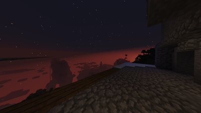 A pixelated landscape setting featuring a sunset or sunrise over blocky terrain with flat surfaces. The sky has a deep red and orange gradient with scattered stars, indicating a nighttime or early morning scene. In the foreground, there is a rocky and cobblestone surface leading towards a silhouetted horizon of blocky mountains and trees.