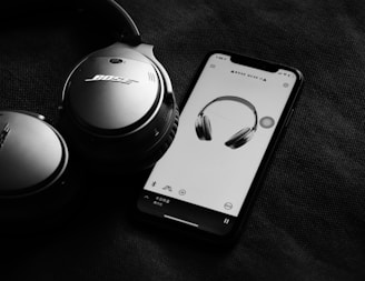 A pair of over-ear headphones is positioned beside a smartphone displaying an image of the same headphones on its screen. The headphones are prominently branded and the phone app appears to be controlling or associated with the headphones. The overall scene is set against a dark fabric background, creating a sleek and sophisticated appearance.