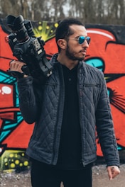 A man in a quilted jacket holds a large video camera over his shoulder. He is wearing aviator sunglasses and stands in front of a colorful graffiti-covered wall with abstract designs.