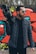 A man in a quilted jacket holds a large video camera over his shoulder. He is wearing aviator sunglasses and stands in front of a colorful graffiti-covered wall with abstract designs.