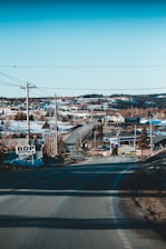 A rural road stretches out towards a small town, flanked by utility poles and power lines. Snow covers parts of the landscape, and several houses are visible in the distance. A few signs are placed along the roadside, with clear skies and hills in the background.