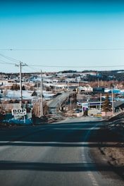 A rural road stretches out towards a small town, flanked by utility poles and power lines. Snow covers parts of the landscape, and several houses are visible in the distance. A few signs are placed along the roadside, with clear skies and hills in the background.