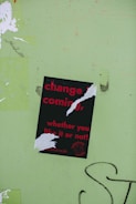 A torn poster with red text on a black background reads 'change is coming, whether you like it or not!' is affixed to a light green wall. The poster has been ripped in several places, partially obscuring some of the words. There is graffiti in black on the wall near the bottom right of the image.