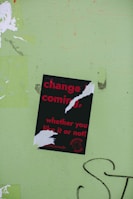 A torn poster with red text on a black background reads 'change is coming, whether you like it or not!' is affixed to a light green wall. The poster has been ripped in several places, partially obscuring some of the words. There is graffiti in black on the wall near the bottom right of the image.