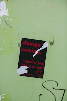 A torn poster with red text on a black background reads 'change is coming, whether you like it or not!' is affixed to a light green wall. The poster has been ripped in several places, partially obscuring some of the words. There is graffiti in black on the wall near the bottom right of the image.
