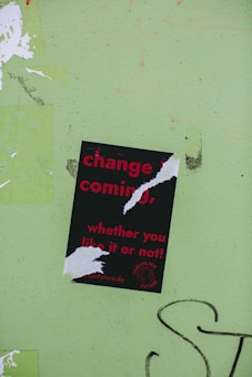 A torn poster with red text on a black background reads 'change is coming, whether you like it or not!' is affixed to a light green wall. The poster has been ripped in several places, partially obscuring some of the words. There is graffiti in black on the wall near the bottom right of the image.