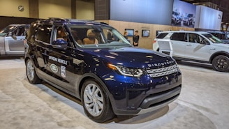 A navy blue Land Rover Discovery is prominently displayed indoors, marked as an official vehicle with various logos on the side. It is surrounded by a spacious, well-lit showroom environment, where other vehicles and display boards can be seen in the background. Two people are standing near another vehicle, possibly staff or attendees.