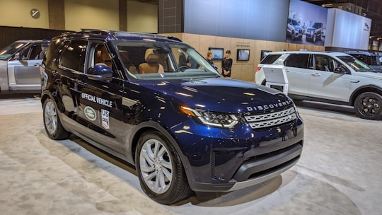 A navy blue Land Rover Discovery is prominently displayed indoors, marked as an official vehicle with various logos on the side. It is surrounded by a spacious, well-lit showroom environment, where other vehicles and display boards can be seen in the background. Two people are standing near another vehicle, possibly staff or attendees.