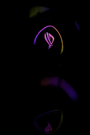 A stylized logo with a futuristic design is illuminated by neon lights in shades of pink, purple, and yellow against a dark background. The curved lines create a sense of motion and technological sophistication.