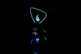 A dark scene with futuristic, neon-lit objects. The central focus is a glowing geometric shape with a stylized logo inside. The neon lights are predominantly blue and green, creating a high-tech atmosphere.