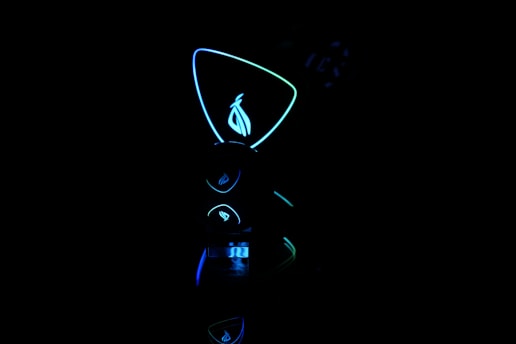 A dark scene with futuristic, neon-lit objects. The central focus is a glowing geometric shape with a stylized logo inside. The neon lights are predominantly blue and green, creating a high-tech atmosphere.